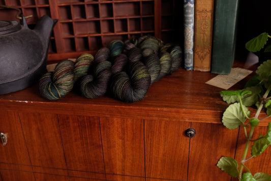 A still life including a cast iron kettle, three green skeins of variegated merino yarn, Swedish Ivy, and three vintage books on top of a vintage Lane cedar chest and in front of a moveable type tray..